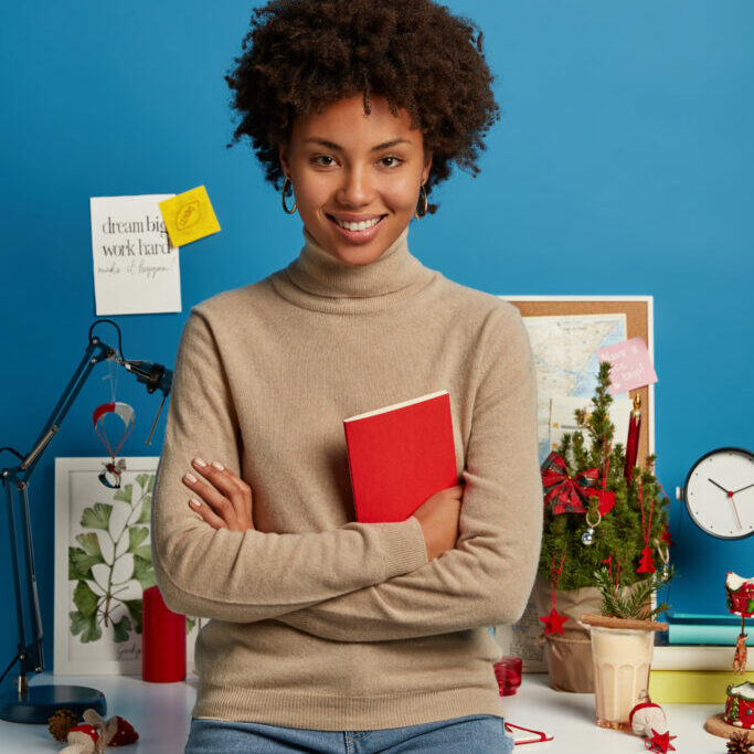 Vertical shot of pleased curly haired female teacher prepares for lessons at home, holds red textbook, poses against desktop, wears turtleneck and jeans, isolated on blue wall. Student in studyroom